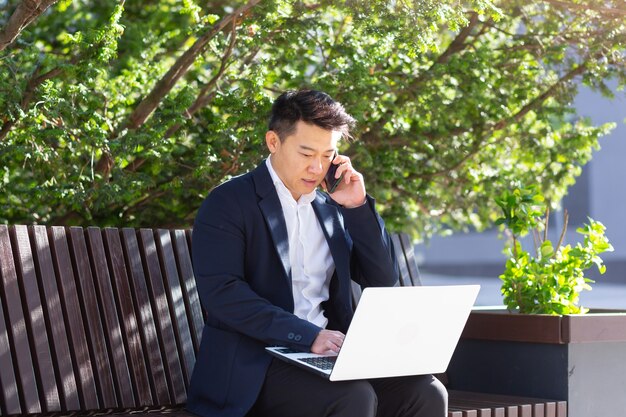 Jovem homem de negócios asiáticos freelancer sentado no banco, trabalhando com o laptop no parque da cidade na rua urbana moderna ao ar livre no centro da cidade.