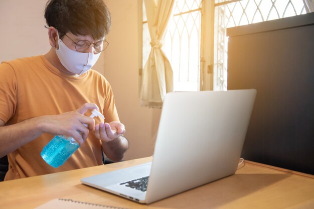 Jovem homem de negócios usando óculos está pressionando o álcool para limpar as mãos, trabalhando no laptop do computador em casa, o evento de crise do vírus corona, covid19