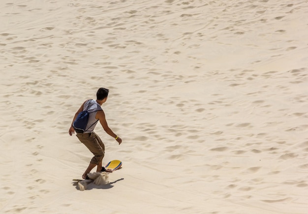 Jovem homem descendo a duna com uma prancha de areia