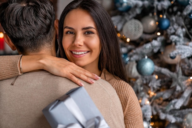 Jovem homem e mulher com uma caixa de presente lindamente decorada abraçando-se perto de uma árvore de Natal