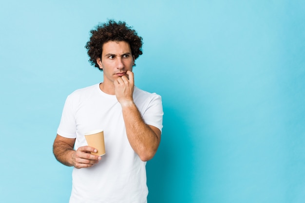 Jovem homem encaracolado caucasiano segurando um café para viagem relaxado pensando em algo olhando para um espaço de cópia.