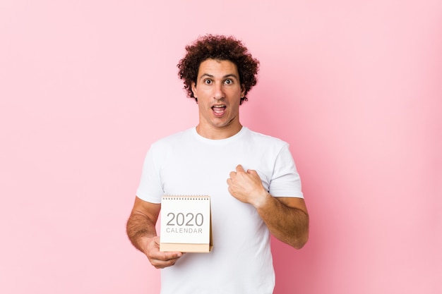 Jovem homem encaracolado caucasiano segurando um calendário 2020 surpreendeu apontando para si mesmo, sorrindo amplamente.