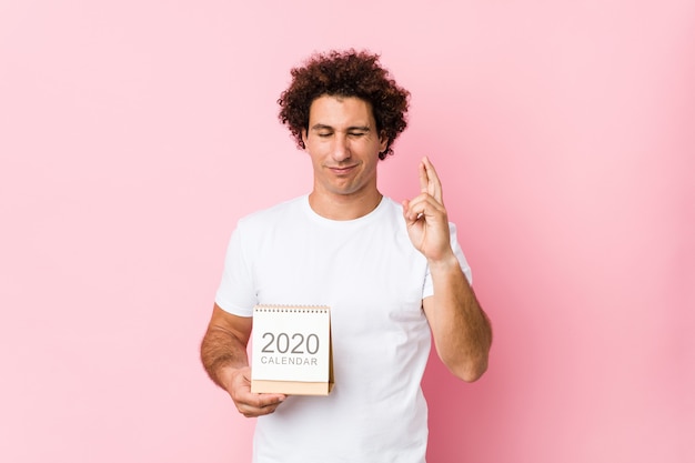 Jovem homem encaracolado caucasiano, segurando um calendário de 2020 cruzar os dedos por ter sorte
