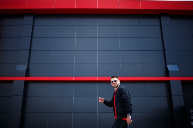 Jovem homem europeu sorridente em casual wear, t-shirt vermelha, jaqueta preta em uma rua da cidade contra um fundo de parede cinza. Cara moderno e estiloso