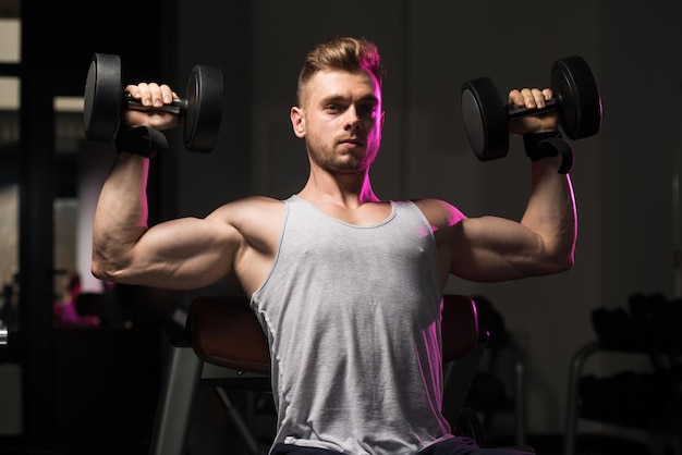 Jovem homem forte na academia e exercitando ombros com halteres Fisiculturista Muscular Atlética Modelo de Aptidão Exercício Ombro