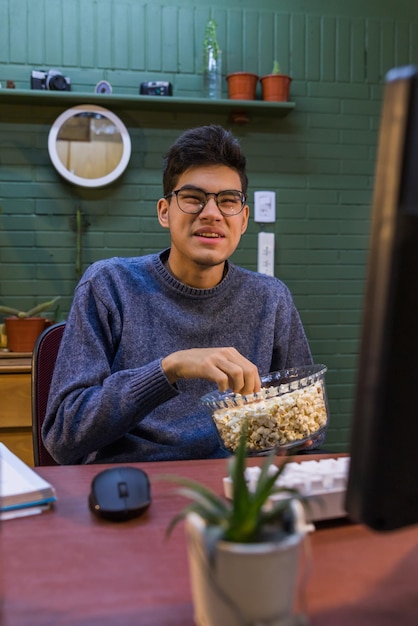 Jovem homem hispânico sorridente sentado em frente ao computador e comendo pipoca