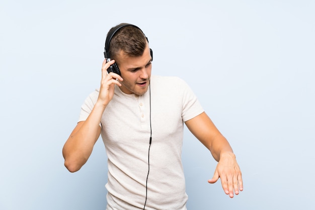 Jovem homem loiro bonito sobre parede azul isolada, ouvindo música com fones de ouvido