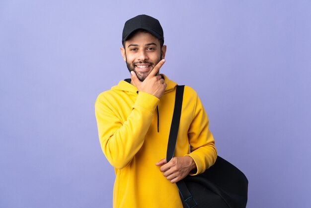 Jovem homem marroquino com bolsa esporte isolada na parede roxa feliz e sorridente