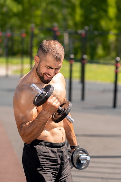 Jovem homem musculoso em topless de short preto se exercitando com halteres pesados em uma manhã ensolarada ao ar livre