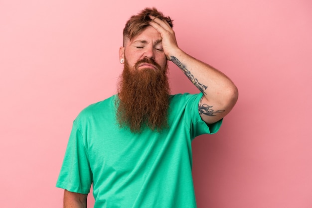 Jovem homem ruivo, caucasiano, com barba comprida, isolado no fundo rosa, cansado e com muito sono, mantendo a mão na cabeça.