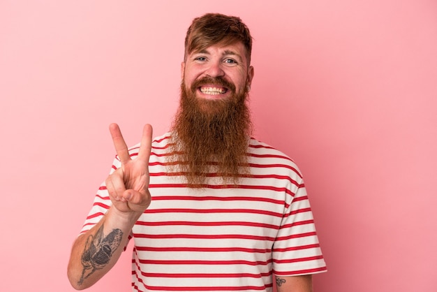 Jovem homem ruivo, caucasiano, com barba comprida, isolado no fundo rosa, mostrando sinal de vitória e sorrindo amplamente.