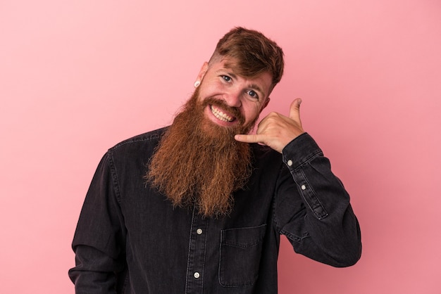 Jovem homem ruivo, caucasiano, com barba comprida, isolado no fundo rosa, mostrando um gesto de chamada de telefone móvel com os dedos.