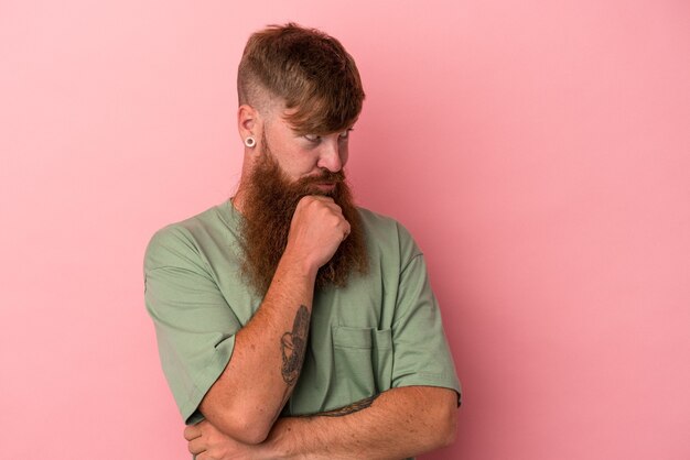 Jovem homem ruivo, caucasiano, com barba comprida, isolado no fundo rosa, olhando de soslaio com expressão duvidosa e cética.