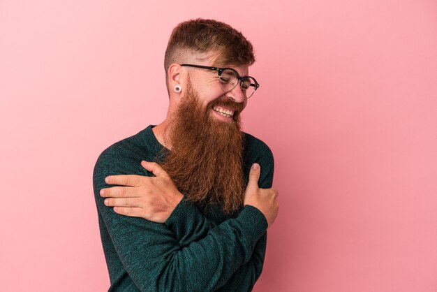 Foto jovem homem ruivo, caucasiano, com barba comprida, isolado no fundo rosa, rindo e se divertindo.