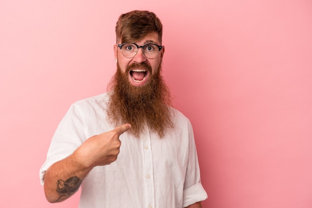 Jovem homem ruivo, caucasiano, com barba comprida, isolado no fundo rosa surpreendeu apontando com o dedo, sorrindo amplamente.