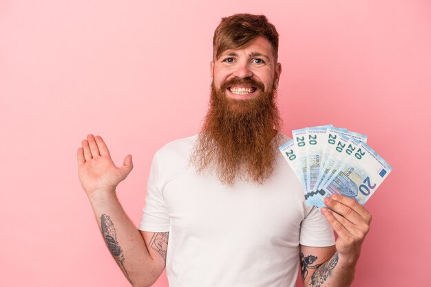 Jovem homem ruivo, caucasiano, com barba comprida, segurando as notas isoladas no fundo rosa, mostrando um espaço de cópia na palma da mão e segurando a outra mão na cintura.