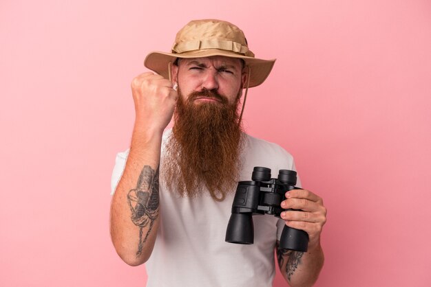 Jovem homem ruivo, caucasiano, com barba comprida, segurando binóculos isolados no fundo rosa, mostrando o punho para a câmera, expressão facial agressiva.