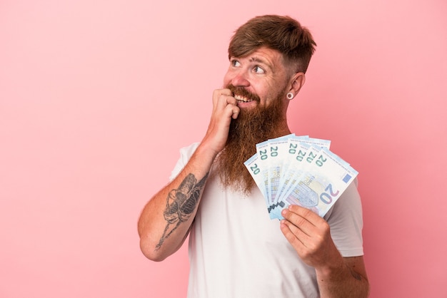 Jovem homem ruivo, caucasiano, com barba comprida, segurando notas isoladas no fundo rosa, relaxado pensando em algo olhando para um espaço de cópia.