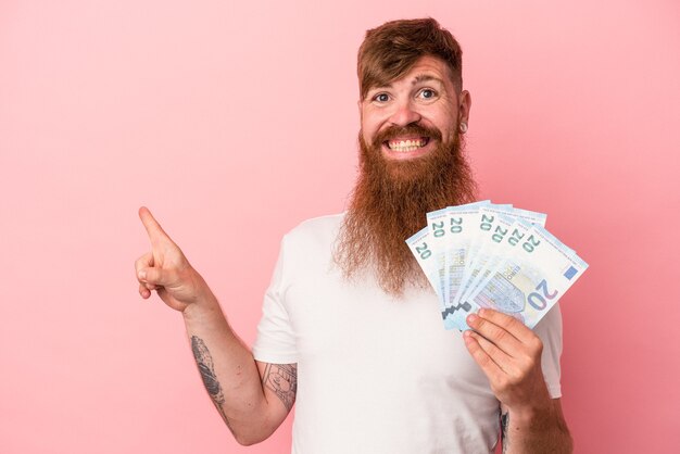 Jovem homem ruivo, caucasiano, com barba comprida, segurando notas isoladas no fundo rosa, sorrindo e apontando de lado, mostrando algo no espaço em branco.