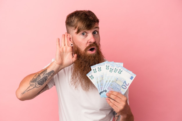 Jovem homem ruivo, caucasiano, com barba comprida, segurando notas isoladas no fundo rosa, tentando ouvir uma fofoca.