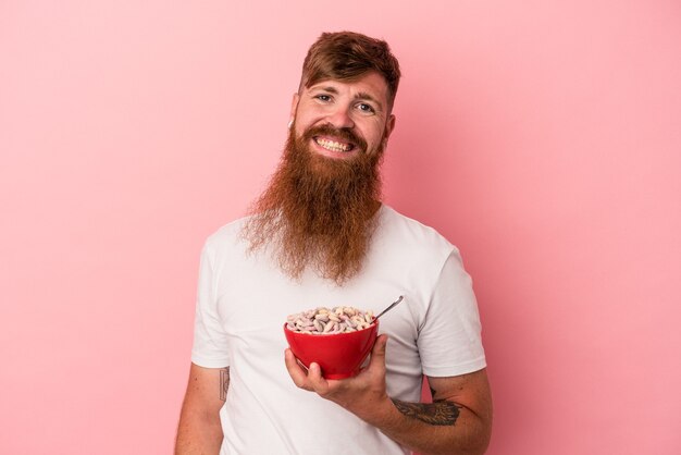 Jovem homem ruivo, caucasiano, com barba comprida, segurando uma tigela de cereales isolada no fundo rosa feliz, sorridente e alegre.