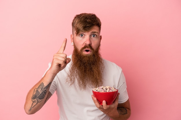 Jovem homem ruivo, caucasiano, com barba comprida, segurando uma tigela de cereales isolada no fundo rosa, tendo uma ideia, o conceito de inspiração.