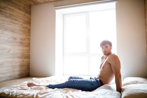 Jovem homem sexy sem camisa na cama no quarto sozinho. Olhe para a câmera e posa. Espera. Homem magro e bem construído, sério, calmo e tranquilo