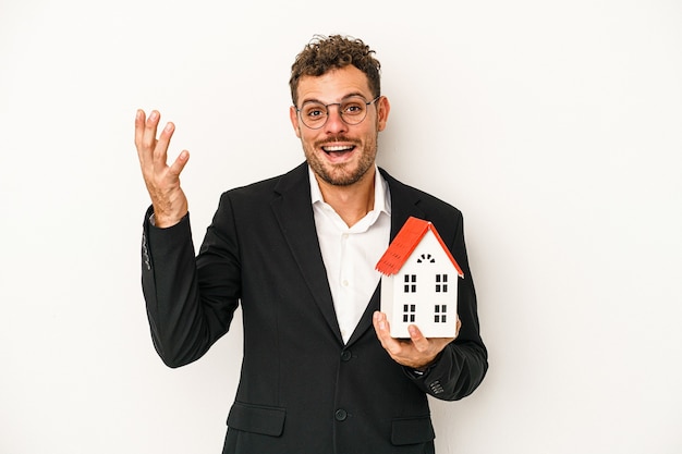 Jovem imobiliário caucasiano segurando uma casa de brinquedo isolada no fundo branco, recebendo uma agradável surpresa, animado e levantando as mãos.