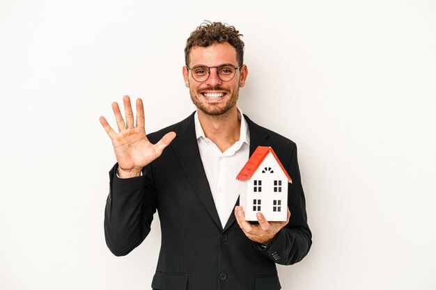 Jovem imobiliário caucasiano segurando uma casa de brinquedo isolada no fundo branco, sorrindo alegre mostrando o número cinco com os dedos.