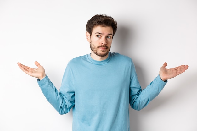 Jovem indeciso com barba, encolhendo os ombros e sem saber de nada, desviando o olhar confuso, em pé de camisa azul sobre fundo branco