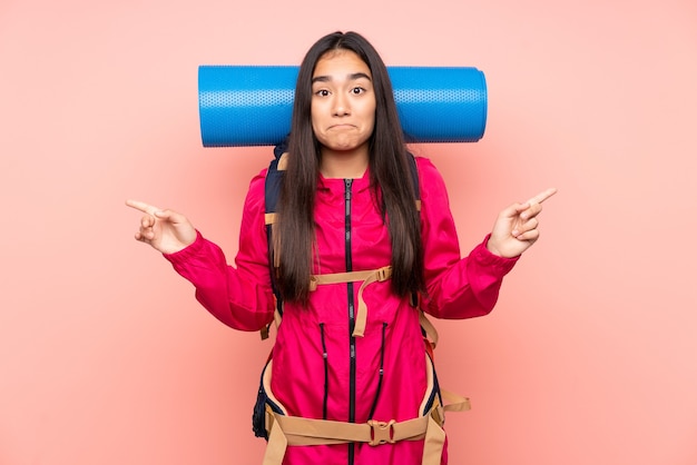 Jovem índia alpinista com uma mochila grande isolada em um fundo rosa apontando para as laterais tendo dúvidas