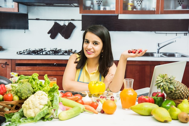 Jovem indiana atraente com avental cozinhando na cozinha com mesa cheia de frutas e vegetais