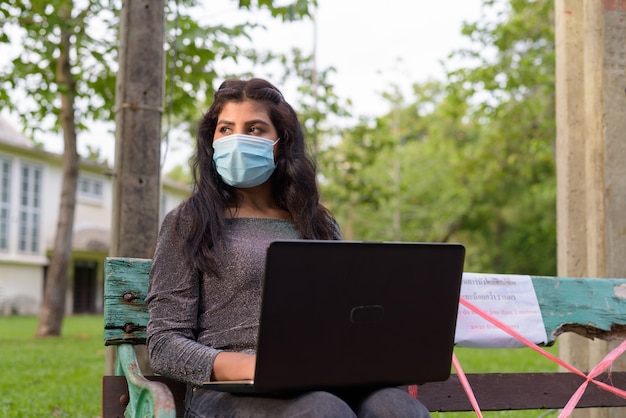 Foto jovem indiana com máscara pensando e usando laptop enquanto está sentada com distância no banco do parque