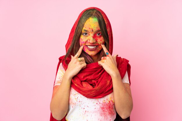 Jovem indiana com pós coloridos de holi no rosto isolado na parede rosa sorrindo com uma expressão feliz e agradável