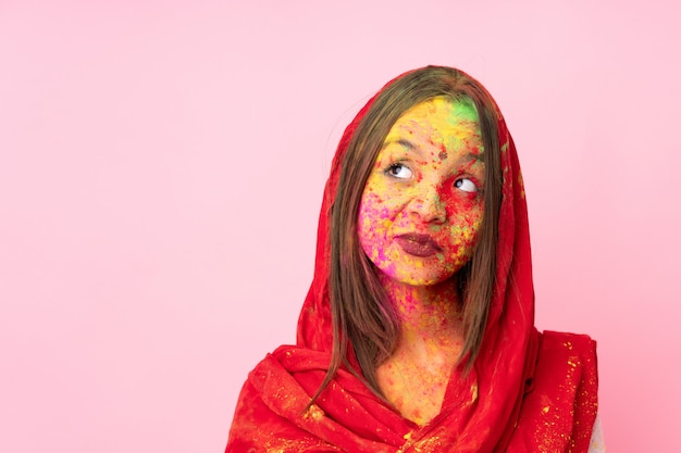 Foto jovem indiana com pós coloridos de holi no rosto na parede rosa com dúvidas enquanto olha de lado
