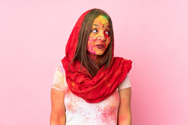 Jovem indiana com pós coloridos de holi no rosto na parede rosa com dúvidas enquanto olha para cima