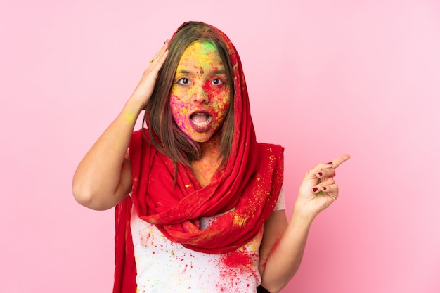 Jovem indiana com pós coloridos de holi no rosto na parede rosa surpreendeu e apontando o dedo para o lado