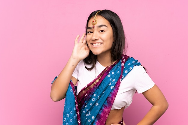 Foto jovem indiana com sari sobre parede ouvindo algo, colocando a mão na orelha