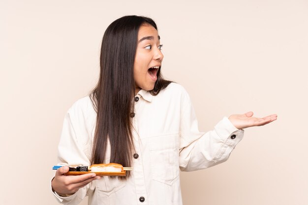 Jovem indiana segurando sushi isolado em um fundo bege com expressão facial de surpresa