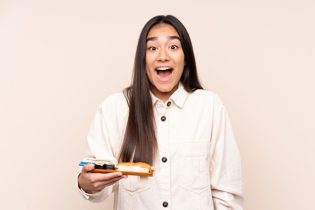 Jovem indiana segurando sushi isolado em um fundo bege com expressão facial surpresa e chocada