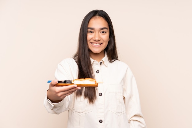 Jovem indiana segurando sushi isolado em um tom bege com uma expressão feliz