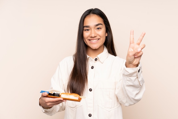 Jovem indiana segurando sushi na parede bege, sorrindo e mostrando sinal de vitória