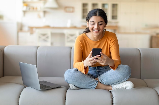 Jovem indiana sorridente usando smartphone e laptop em casa