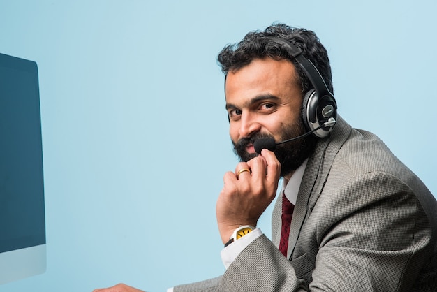 Jovem indiano asiático barbudo de terno em uma central de atendimento, em ambientes internos, ouvindo fones de ouvido, navegando no computador ou fazendo uma chamada de voz