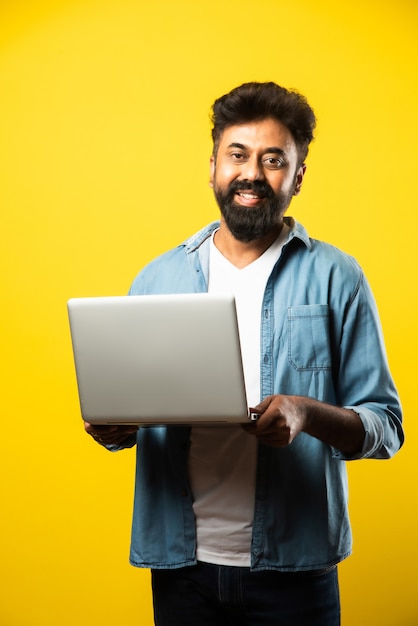 Jovem indiano asiático barbudo em roupas casuais usando laptop em pé no amarelo