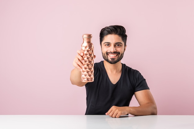 Jovem indiano asiático barbudo segurando ou apresentando uma garrafa de água de cobre contra um fundo rosa enquanto está sentado à mesa