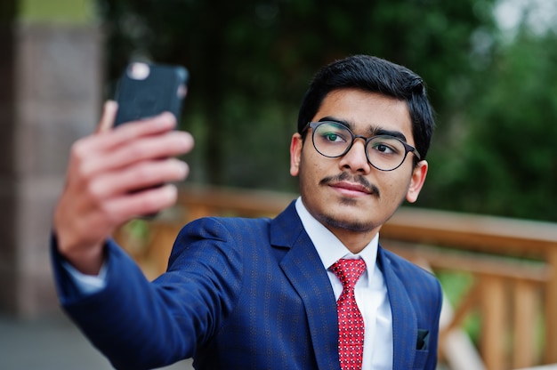Jovem indiano de óculos, vestir terno com gravata vermelha posou ao ar livre e fazendo selfie no celular.