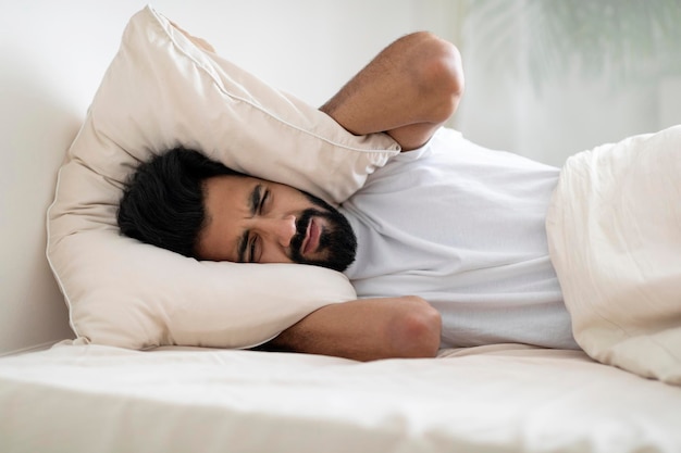 Foto jovem indiano irritado deitado na cama e cobrindo as orelhas com um travesseiro