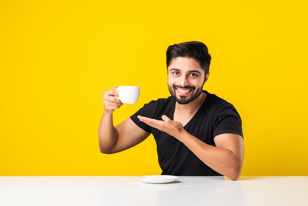Jovem indiano segurando uma xícara de chá, bebendo e saboreando o aroma do café. Homem asiático sentado à mesa contra um fundo amarelo com uma xícara