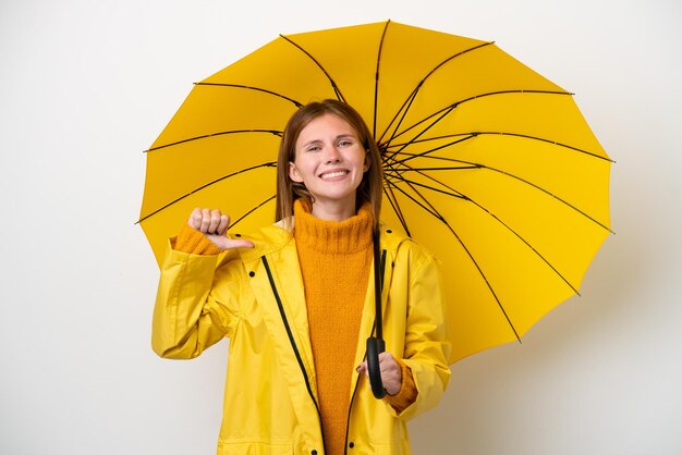 Jovem inglesa com casaco à prova de chuva e guarda-chuva isolado em fundo branco orgulhoso e satisfeito